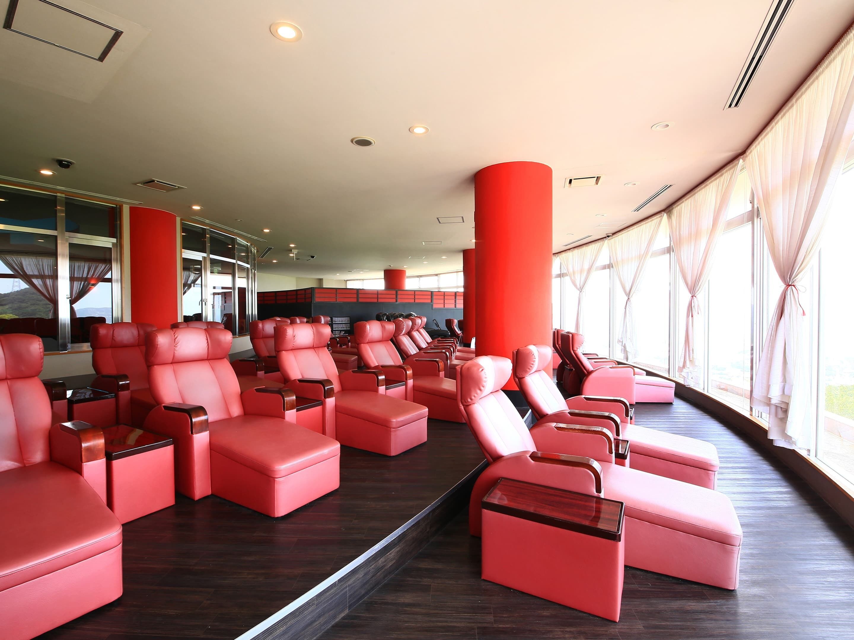 Un área de descanso moderna con filas de sillones y otomanas de cuero rojo, ventanas amplias con cortinas y un techo alto con iluminación empotrada.