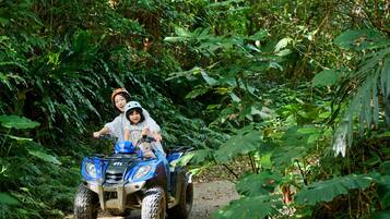 Een blauwe quad met twee personen die over een stoffig pad rijden, omringd door dichte groene beplanting.