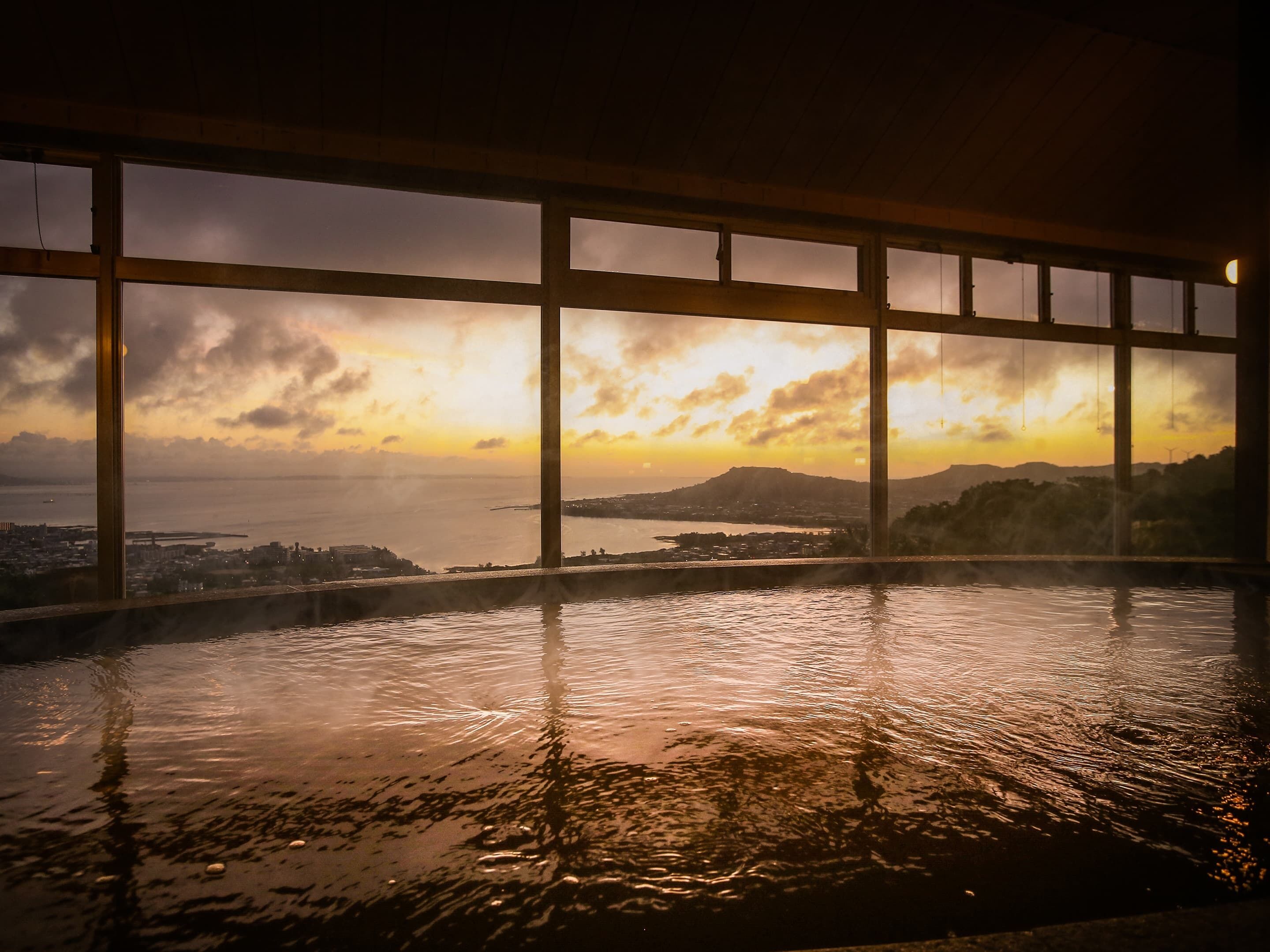A view from an indoor pool area with large windows overlooking a sunset over a body of water and distant land.