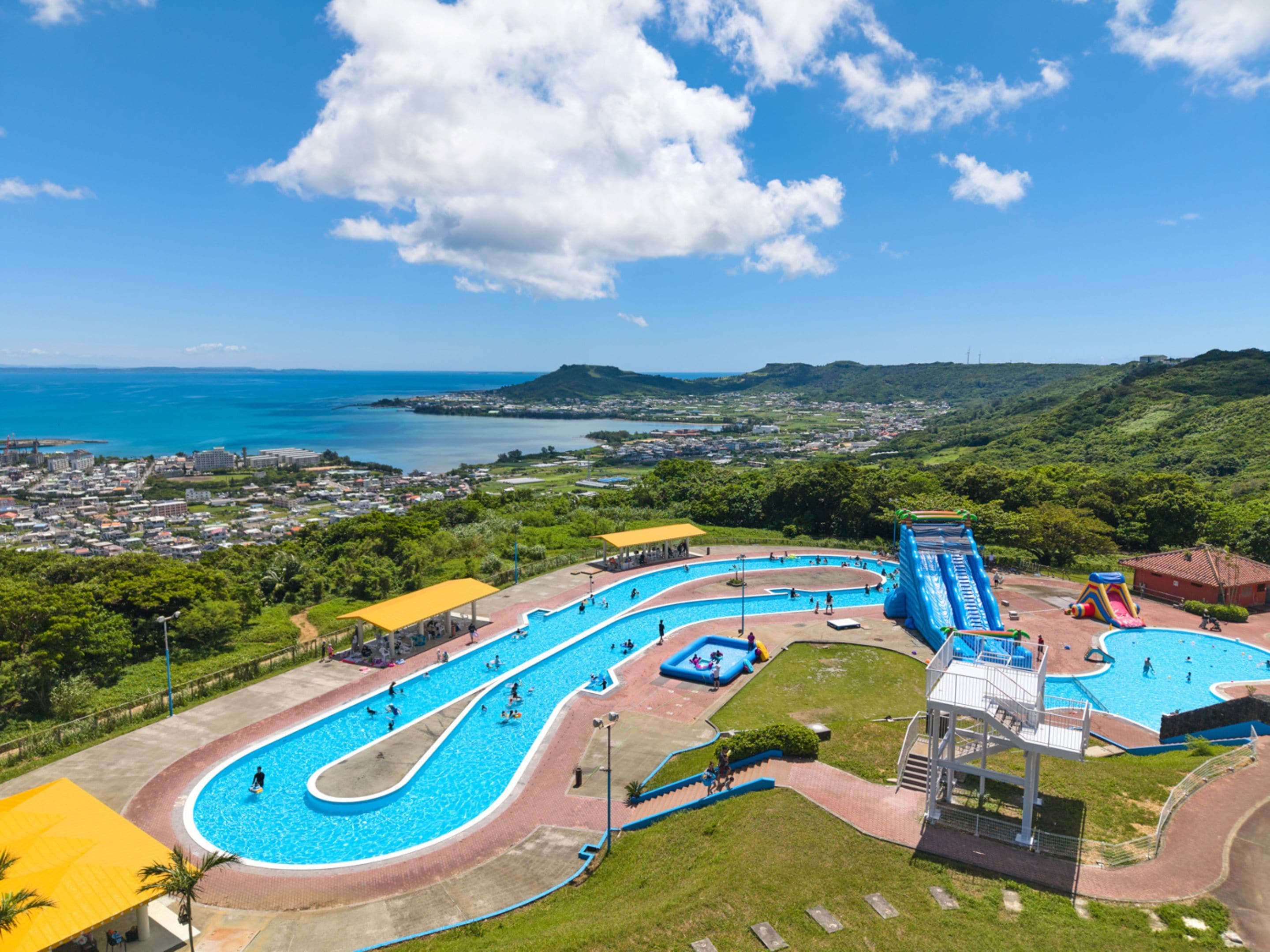 Ein Wasserpark mit mehreren Schwimmbecken und Rutschen, umgeben von viel Grün und mit Blick auf eine Küstenstadt.