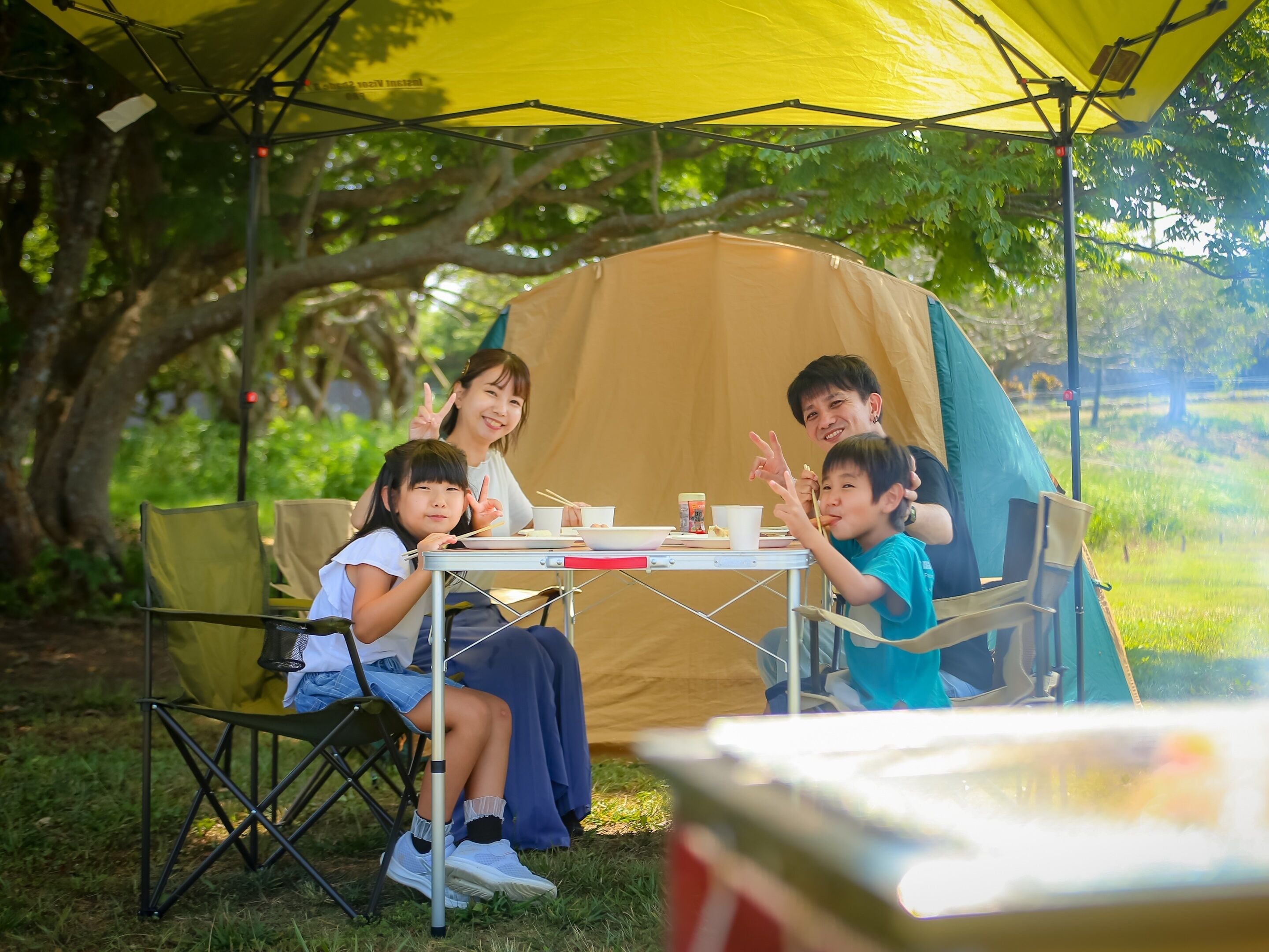A group of people sitting at a table under a yellow canopy, with a tent in the background.