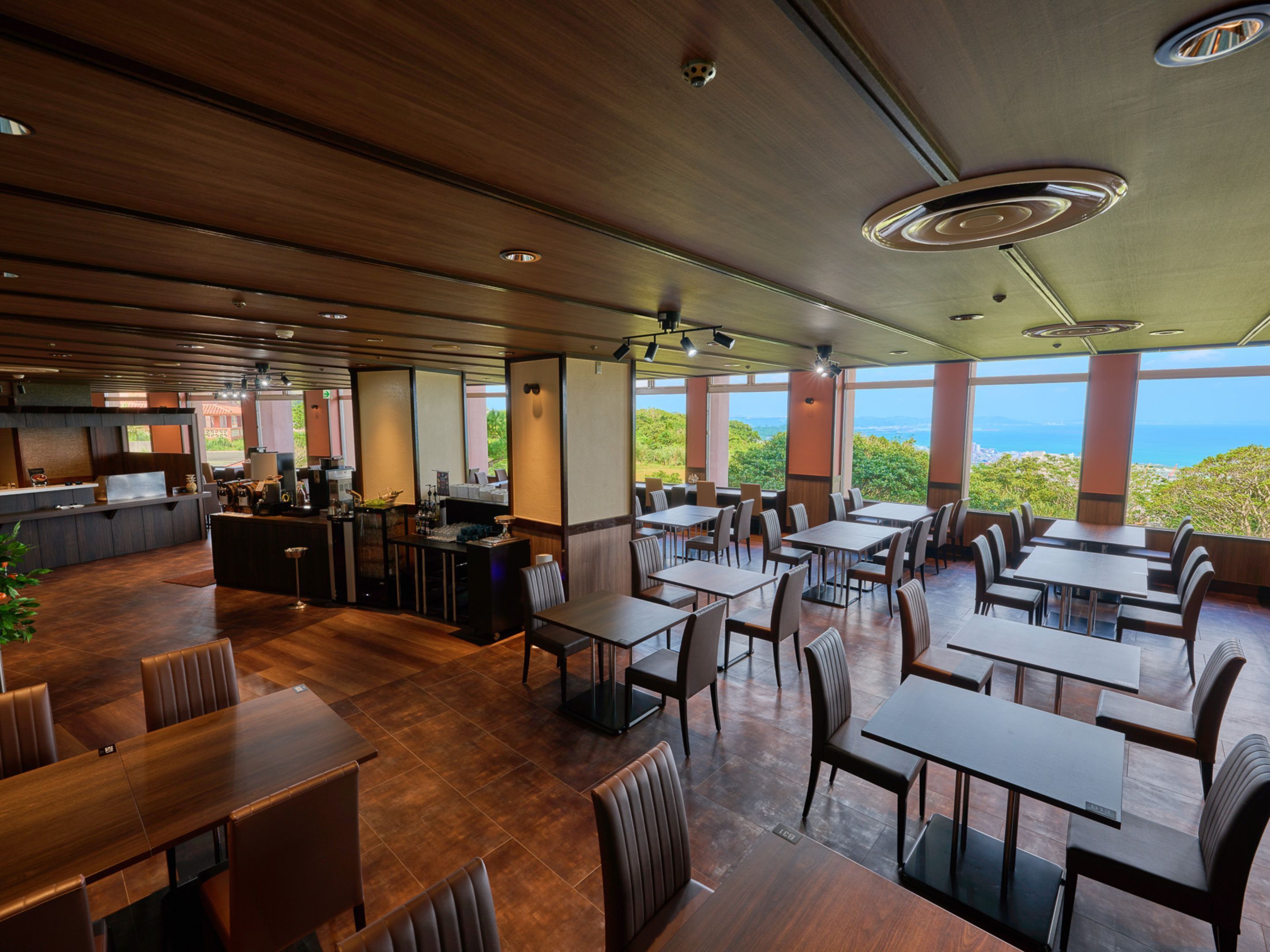 A spacious restaurant with multiple dining tables and chairs, a bar area, and large windows offering a view of the outdoors.