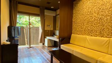 A hotel room with a sofa, a television on a stand, a wooden floor, a stone wall, and a window with curtains.