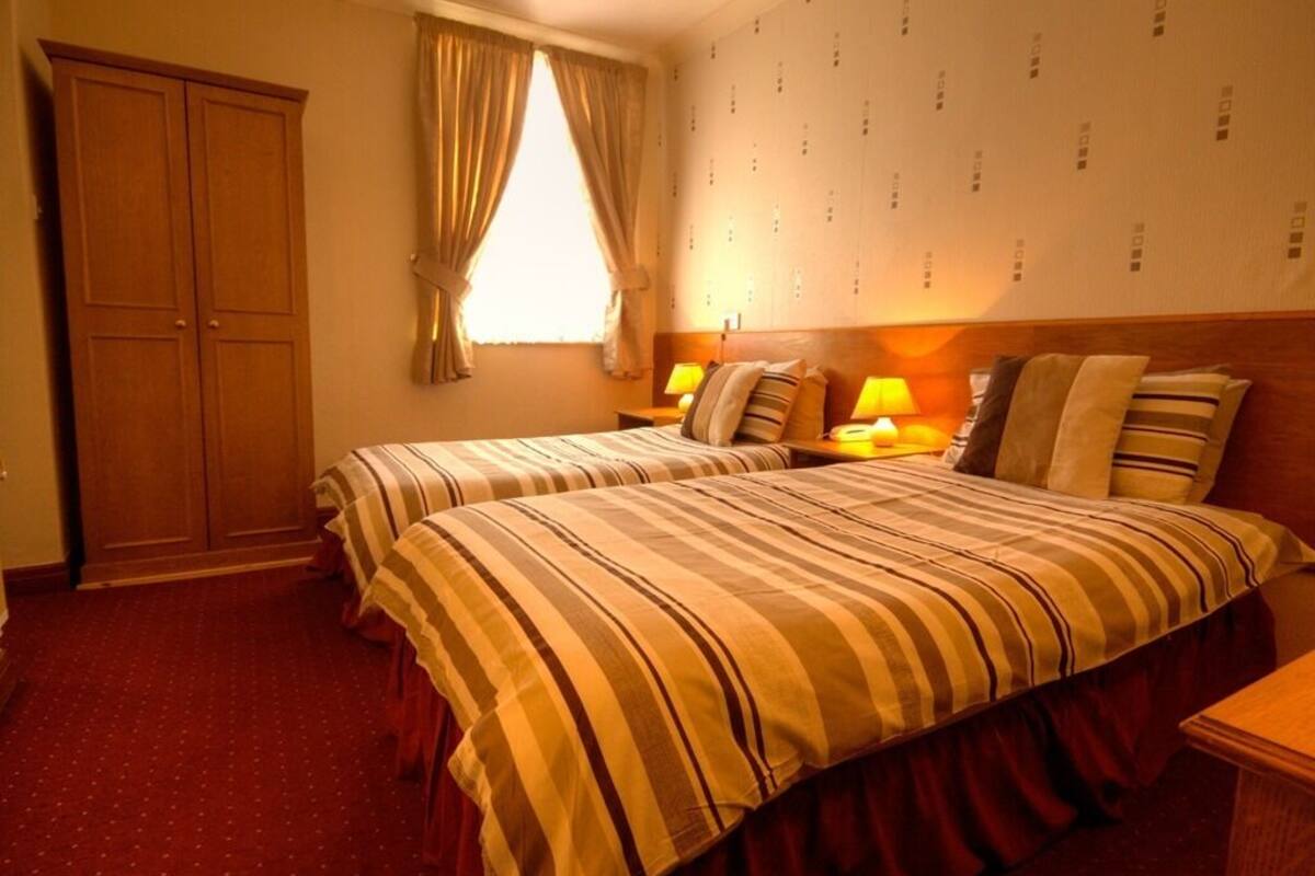 Quarto de hotel com duas camas, um guarda-roupa, uma janela com cortinas e uma parede com padrão.