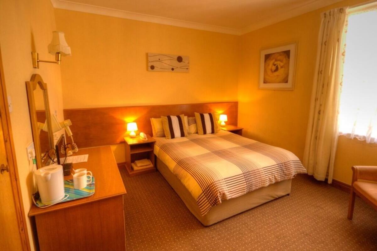 Quarto de hotel com cama, mesas de cabeceira, escrivaninha com luminária, espelho e janela com cortinas.