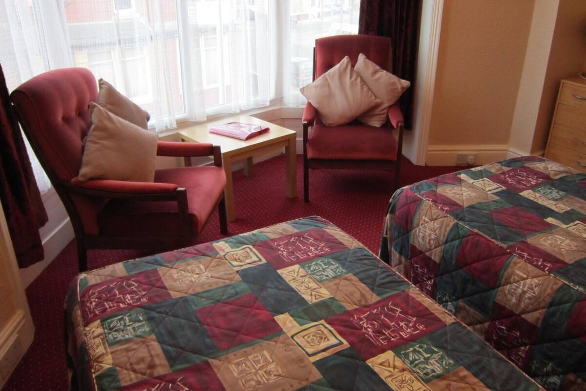 Une chambre d’hôtel avec deux lits, un fauteuil rouge, une petite table et une fenêtre avec des rideaux légers.