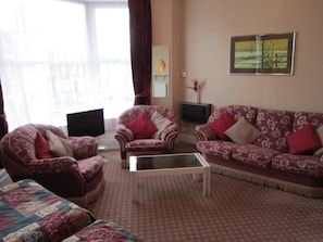 Ein Wohnzimmer mit zwei gemusterten Sesseln, einem Sofa, einem Glastisch und einem Fernseher.
