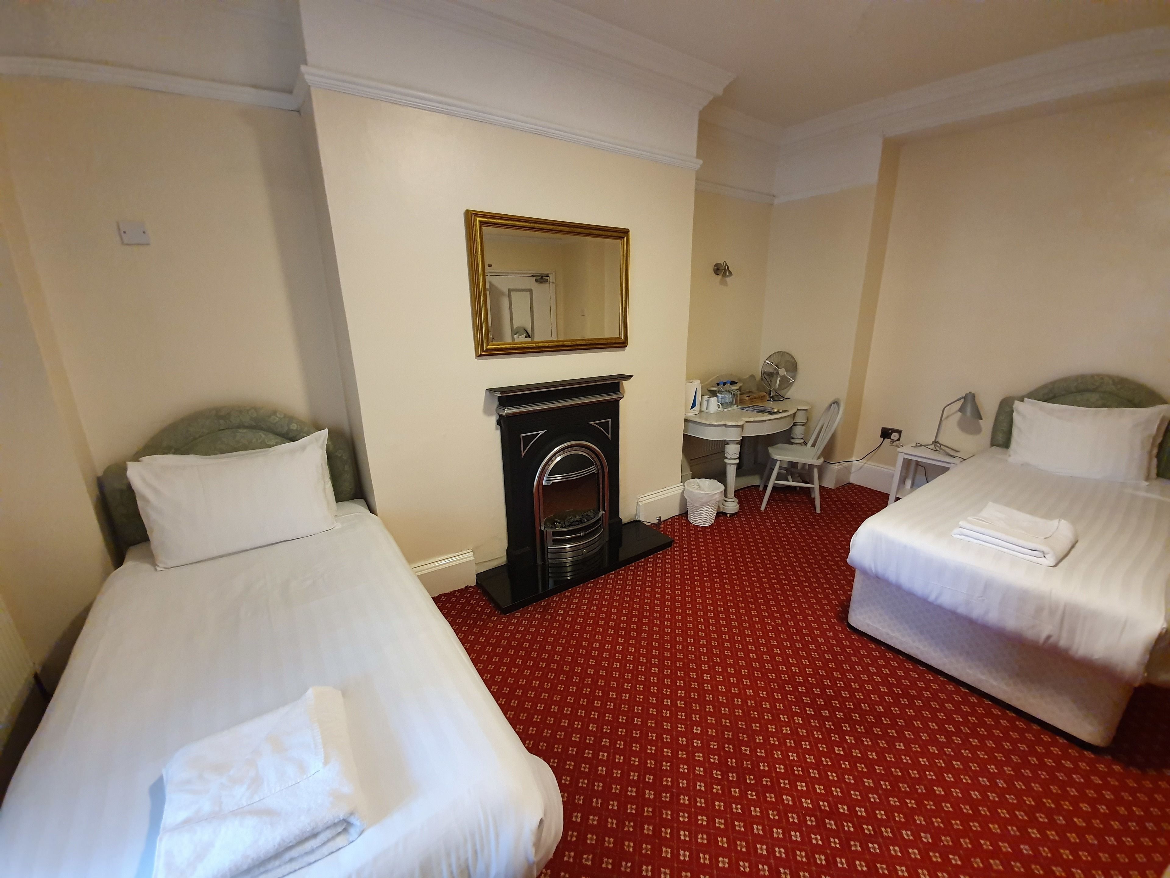 Habitación de hotel con dos camas, un escritorio, una silla, un espejo, una chimenea y una alfombra roja estampada.