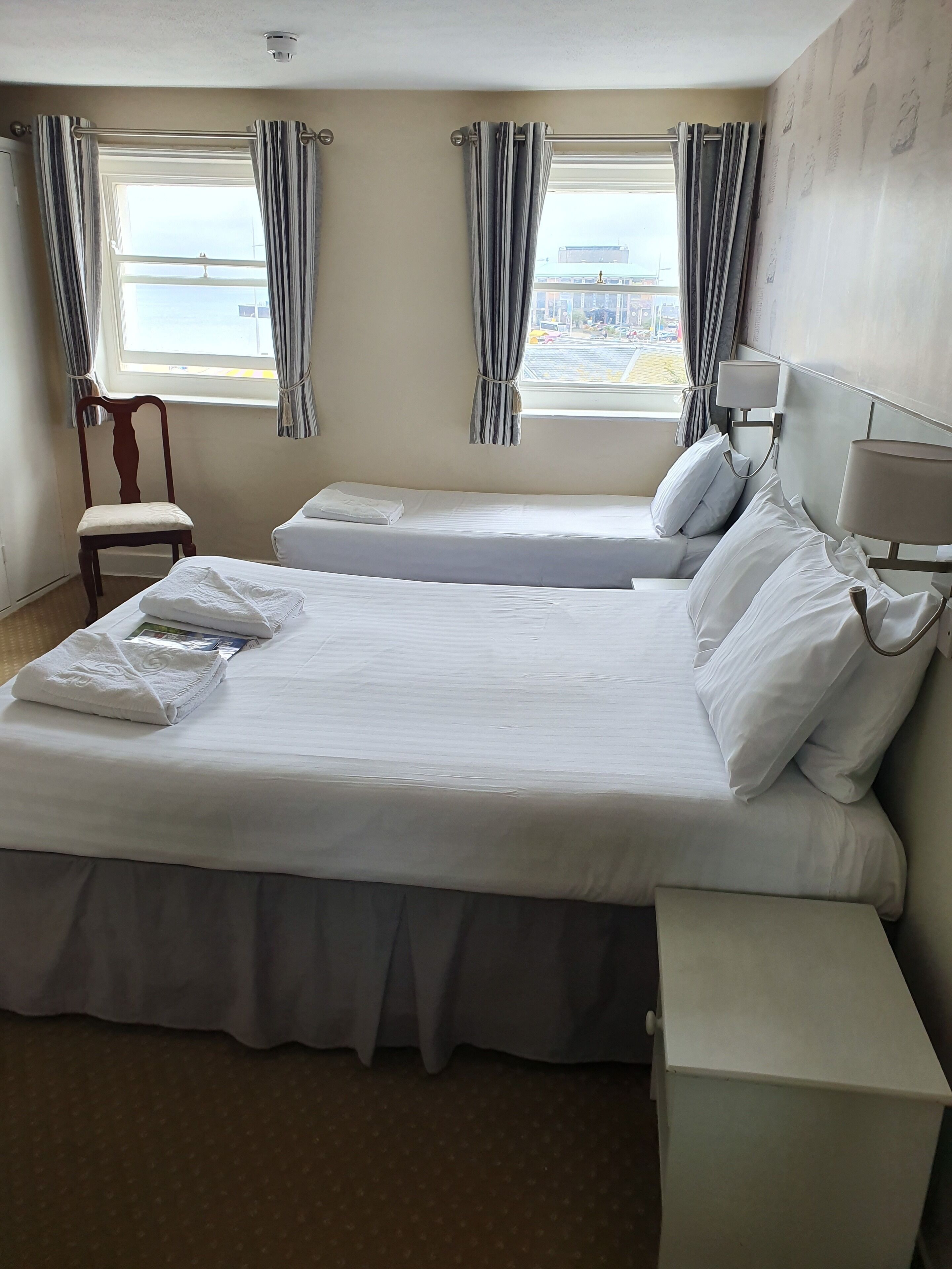 A hotel room with two beds, a chair, a bedside table, and a window with curtains.