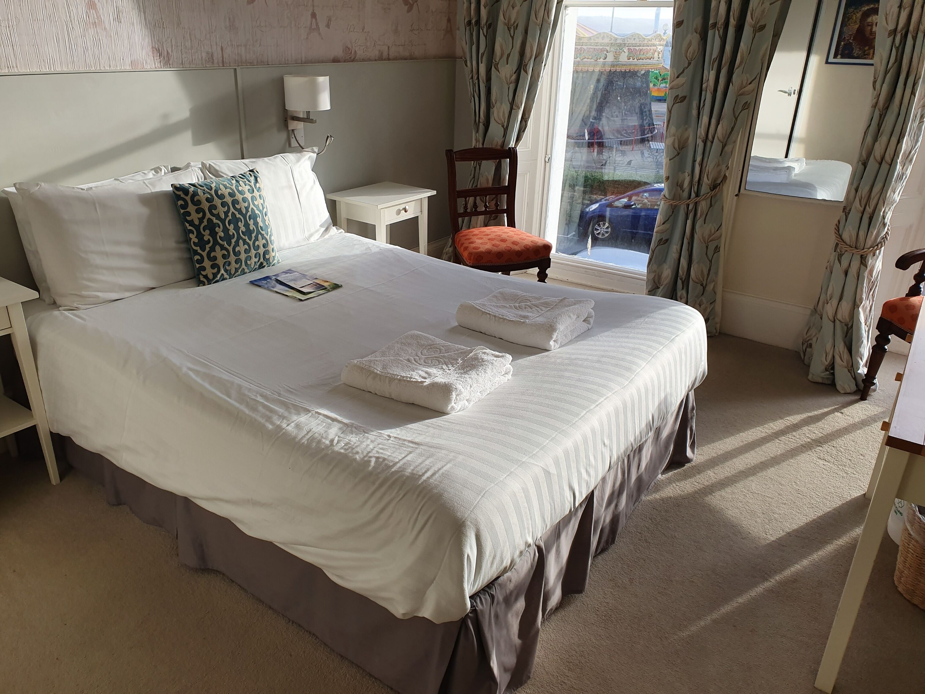 A neatly made bed with white linens and two folded towels, a small bedside table with a lamp, a chair, and a window with curtains.