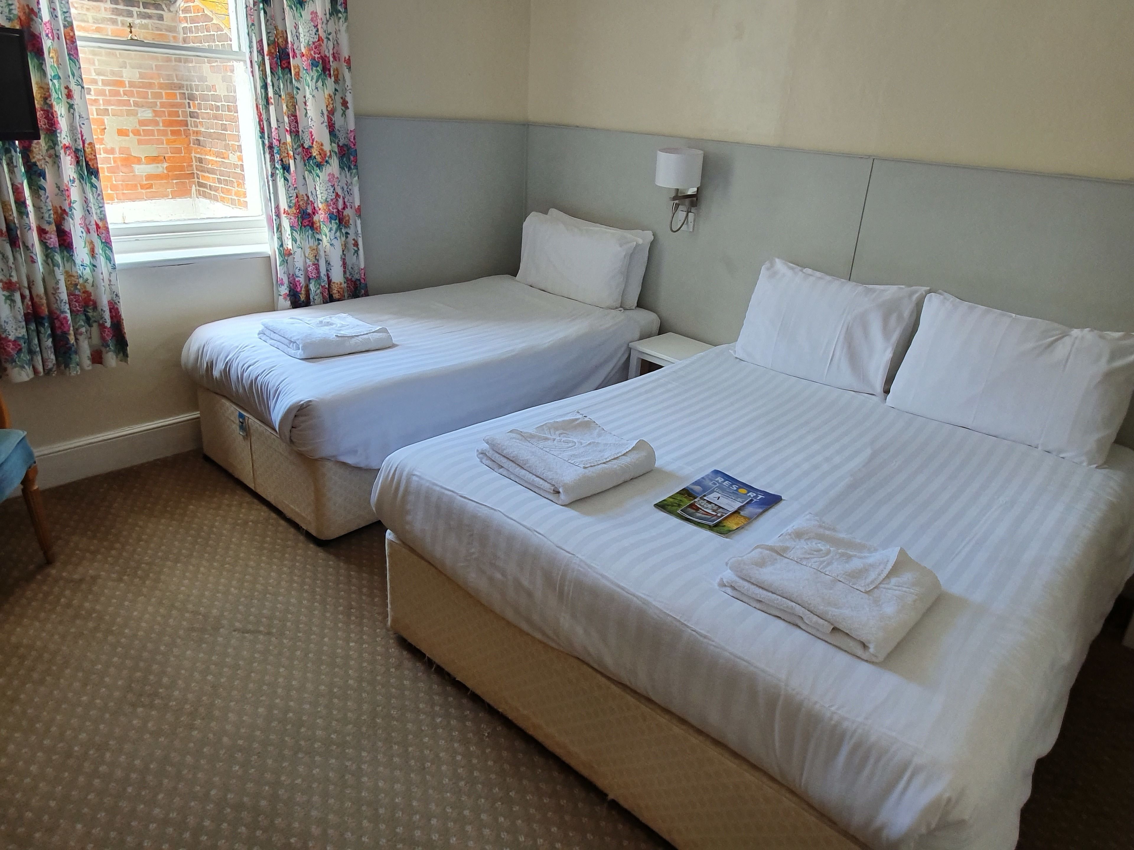 A hotel room with two single beds, a small bedside table, a wall-mounted lamp, and a window with floral curtains.
