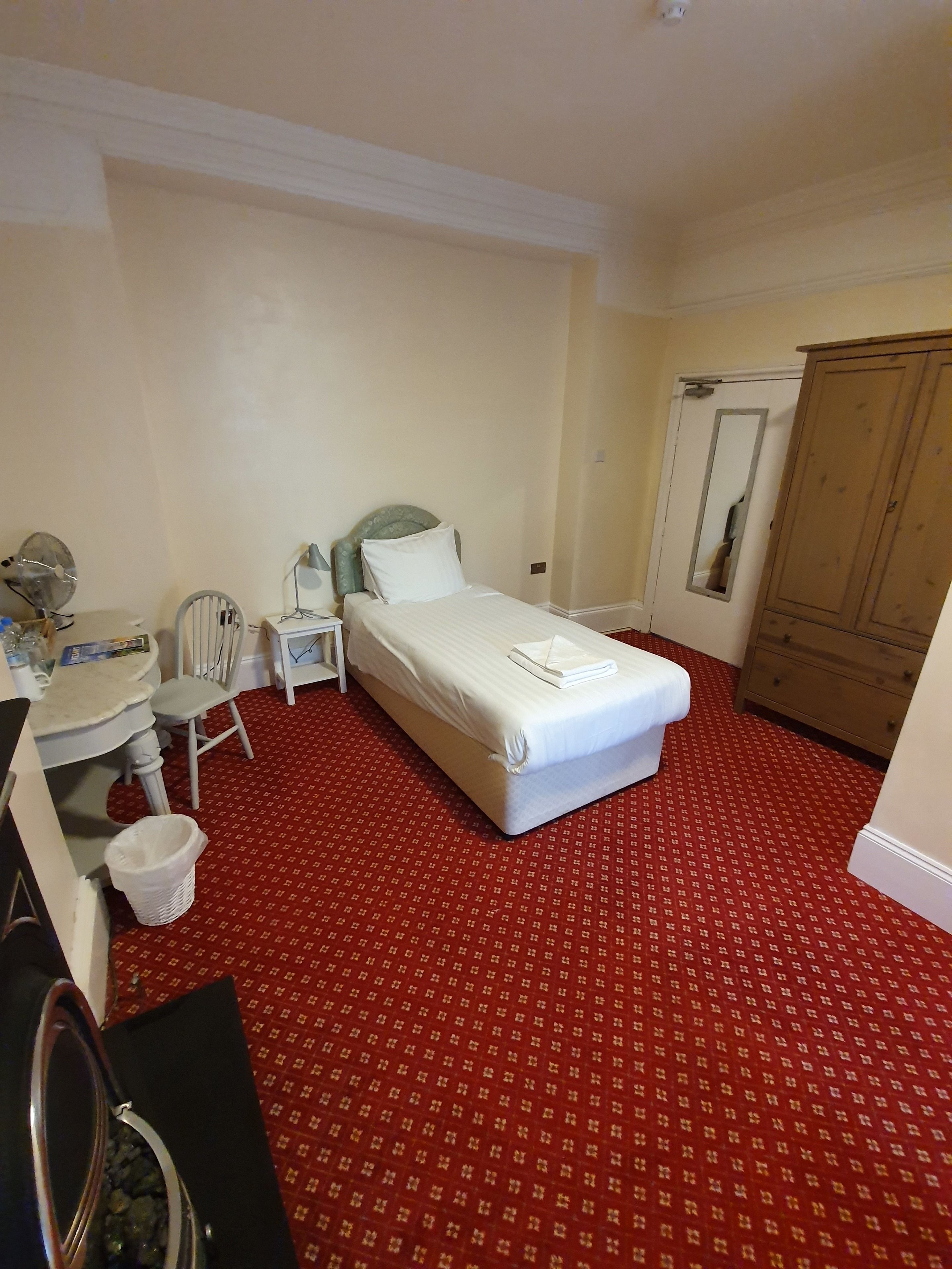 A hotel room with a bed, a desk with a chair, a mirror, a wardrobe, and a red patterned carpet.