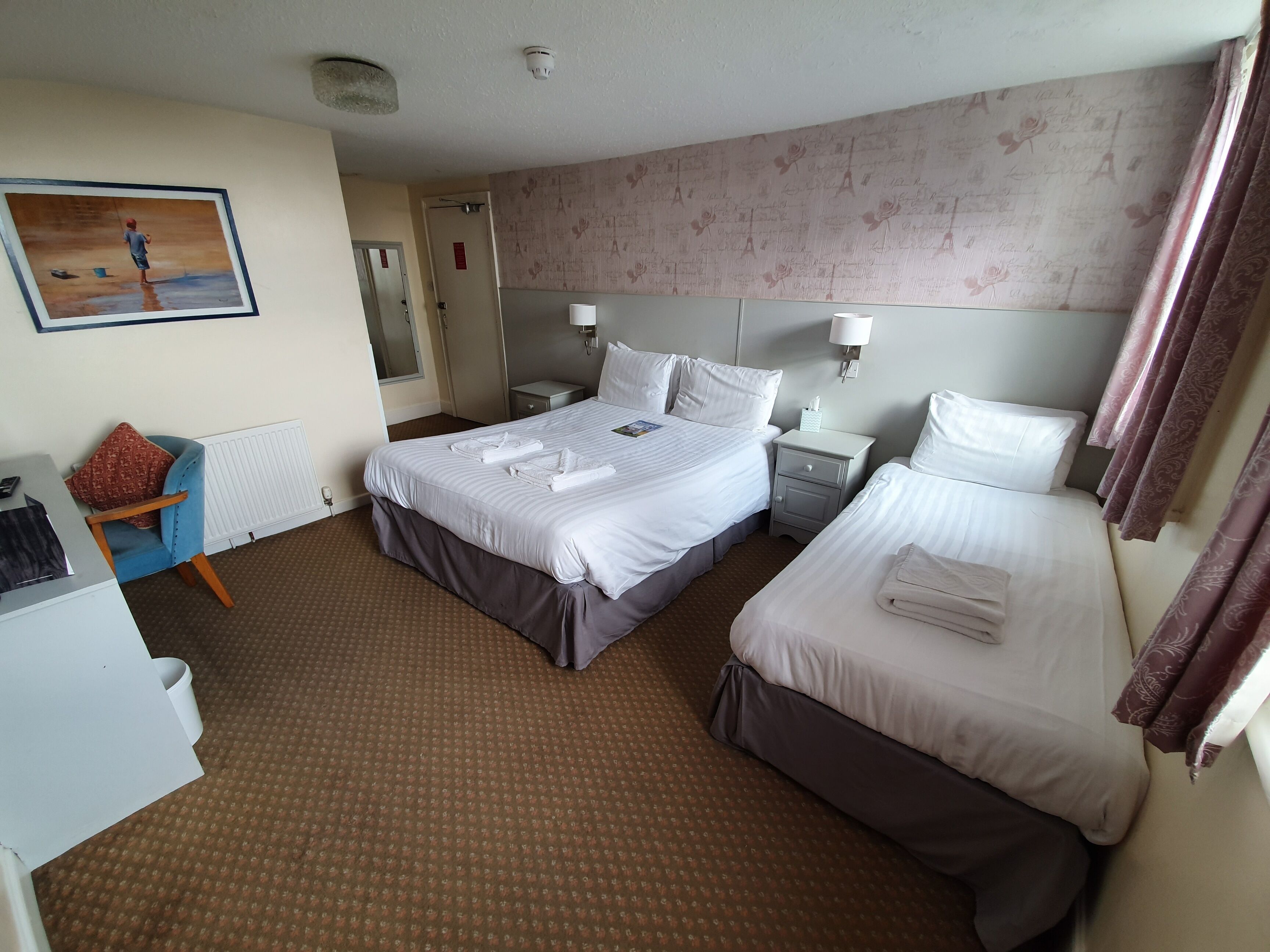 A hotel room with two beds, a desk, a chair, a radiator, a painting, and a nightstand with a lamp.