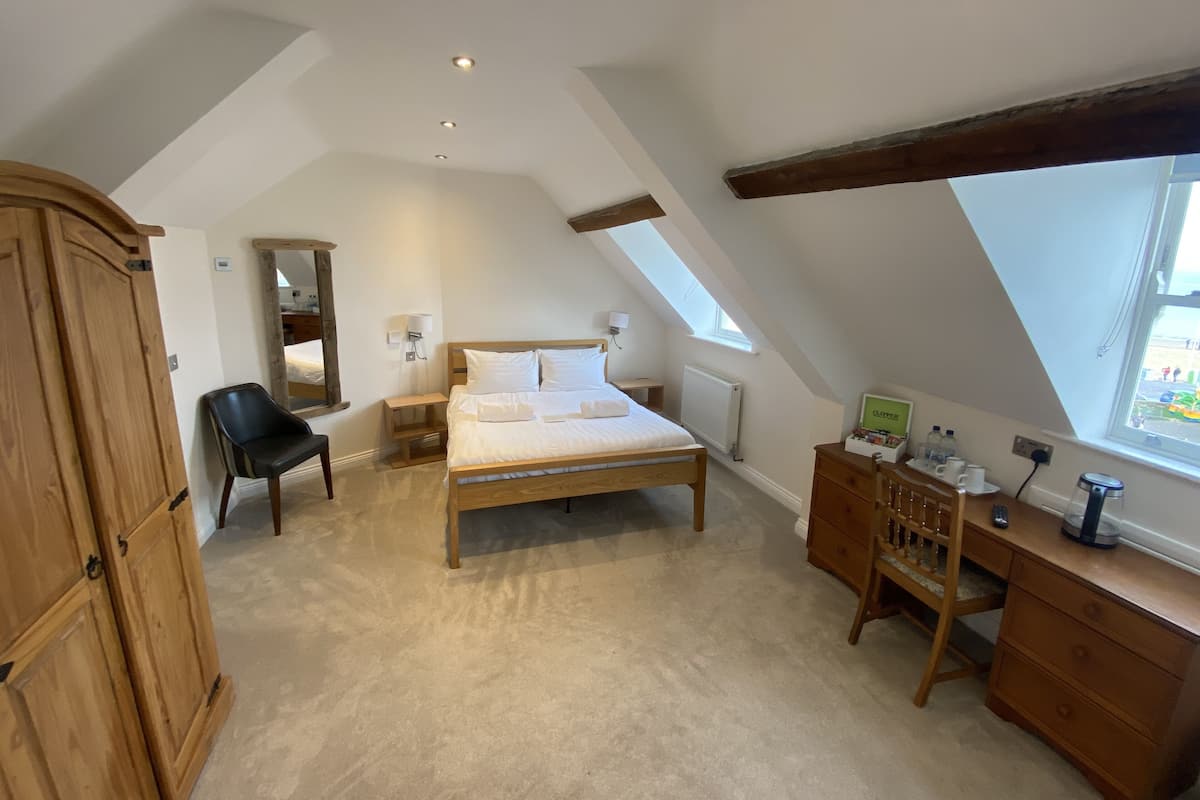 A bedroom with a sloped ceiling, a wooden bed, a desk with a chair, a mirror, and a window.
