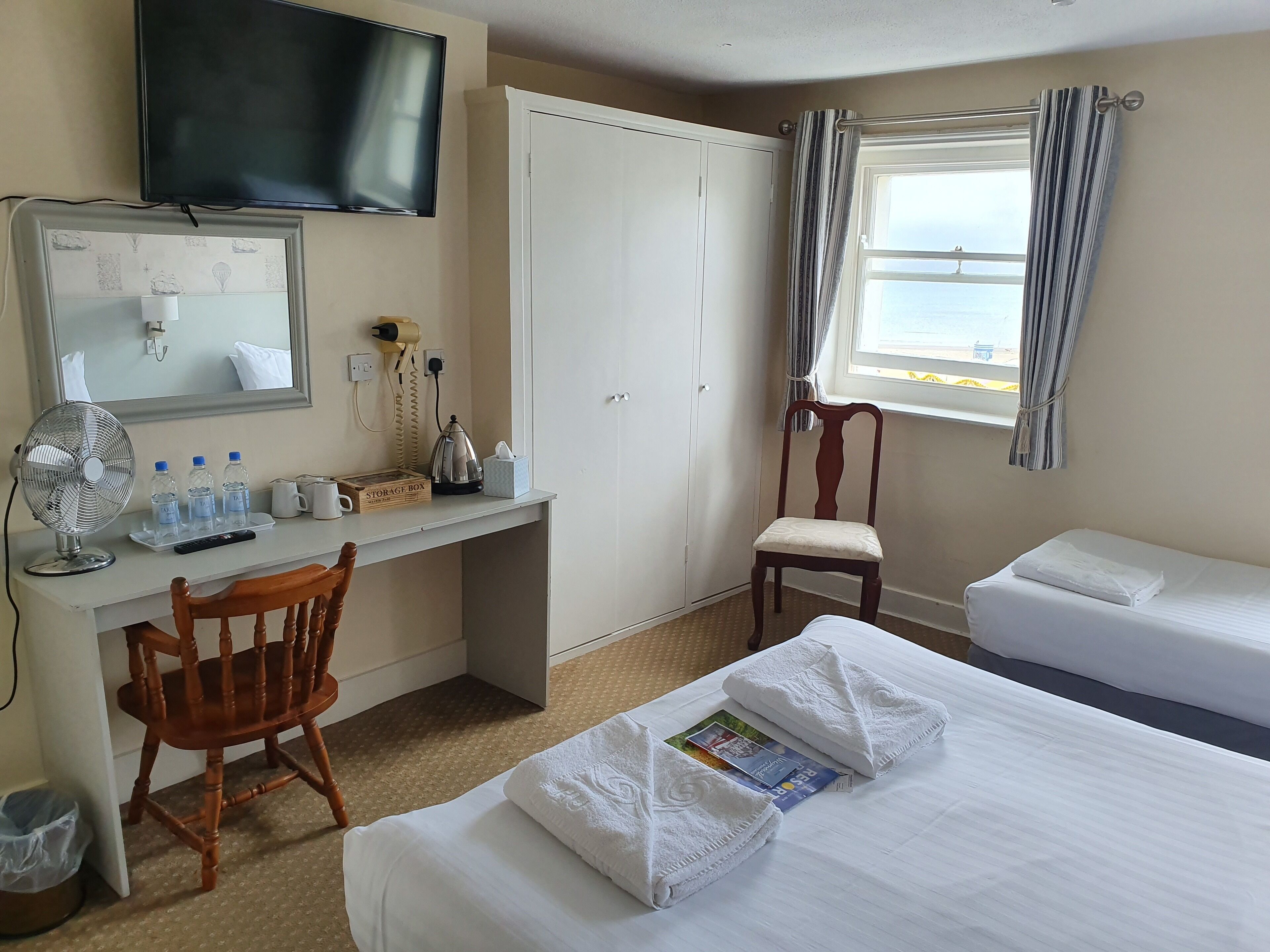 Quarto de hotel com cama, cadeira, escrivaninha com ventoinha, chaleira e caixa organizadora.