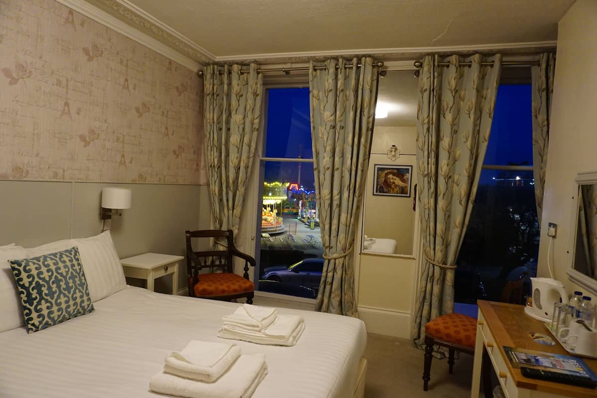 A hotel room with a bed, a chair, a table with a kettle, and a window with curtains.