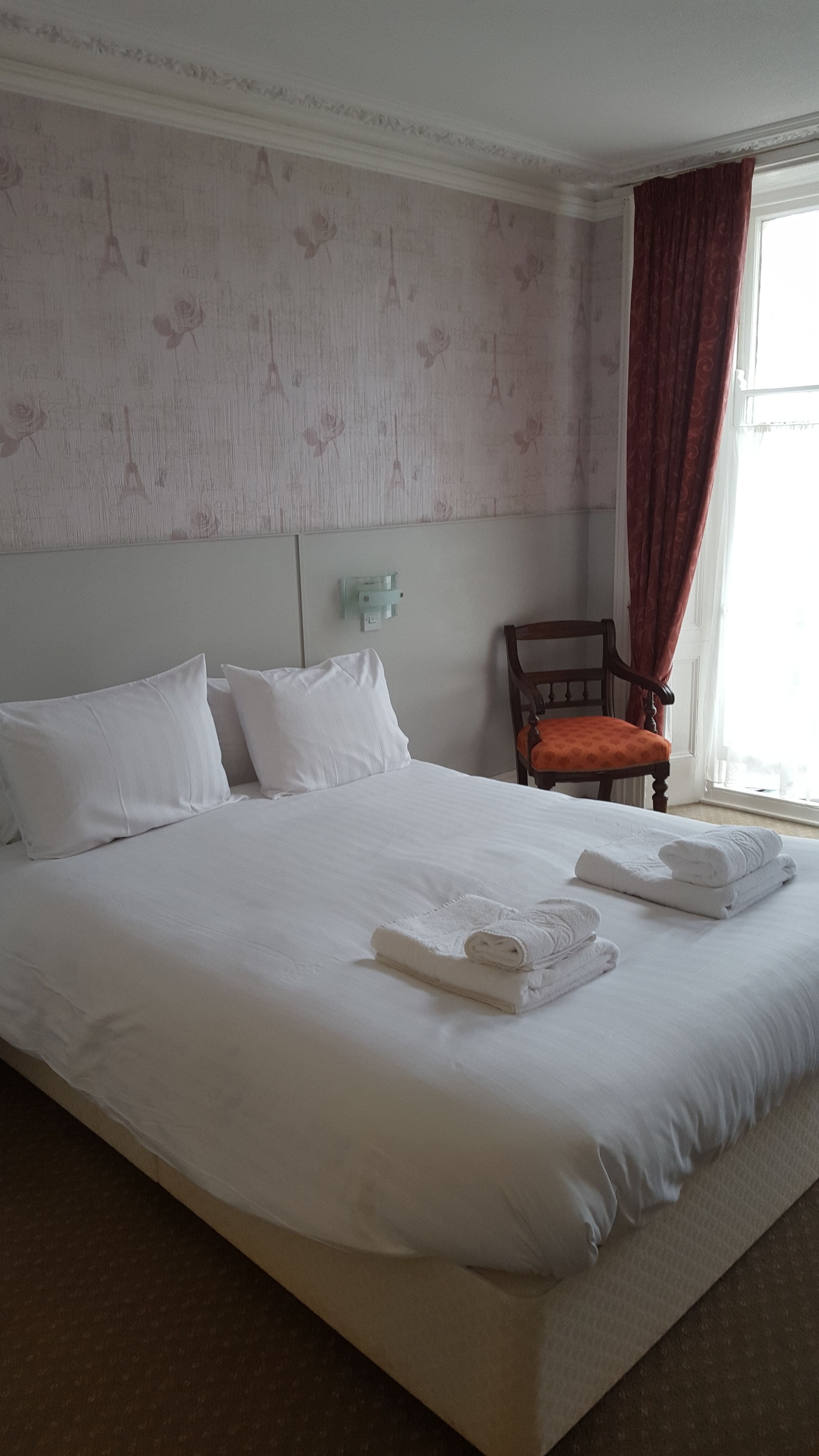 A hotel room with a large bed, white linens, a wooden chair, and a window with curtains.