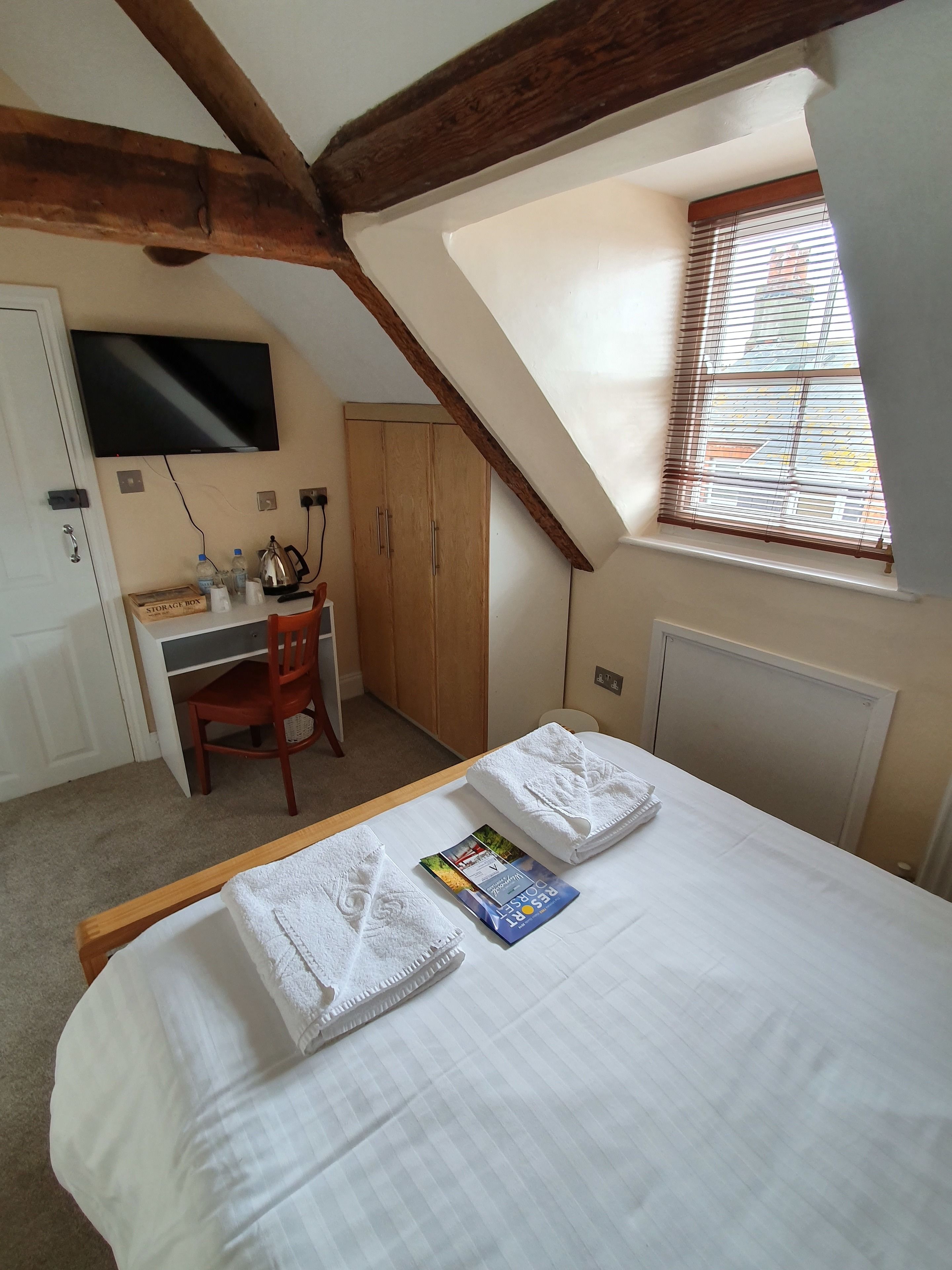 Een slaapkamer met een bed, een bureau, een stoel, een televisie en een raam met jaloezieën.