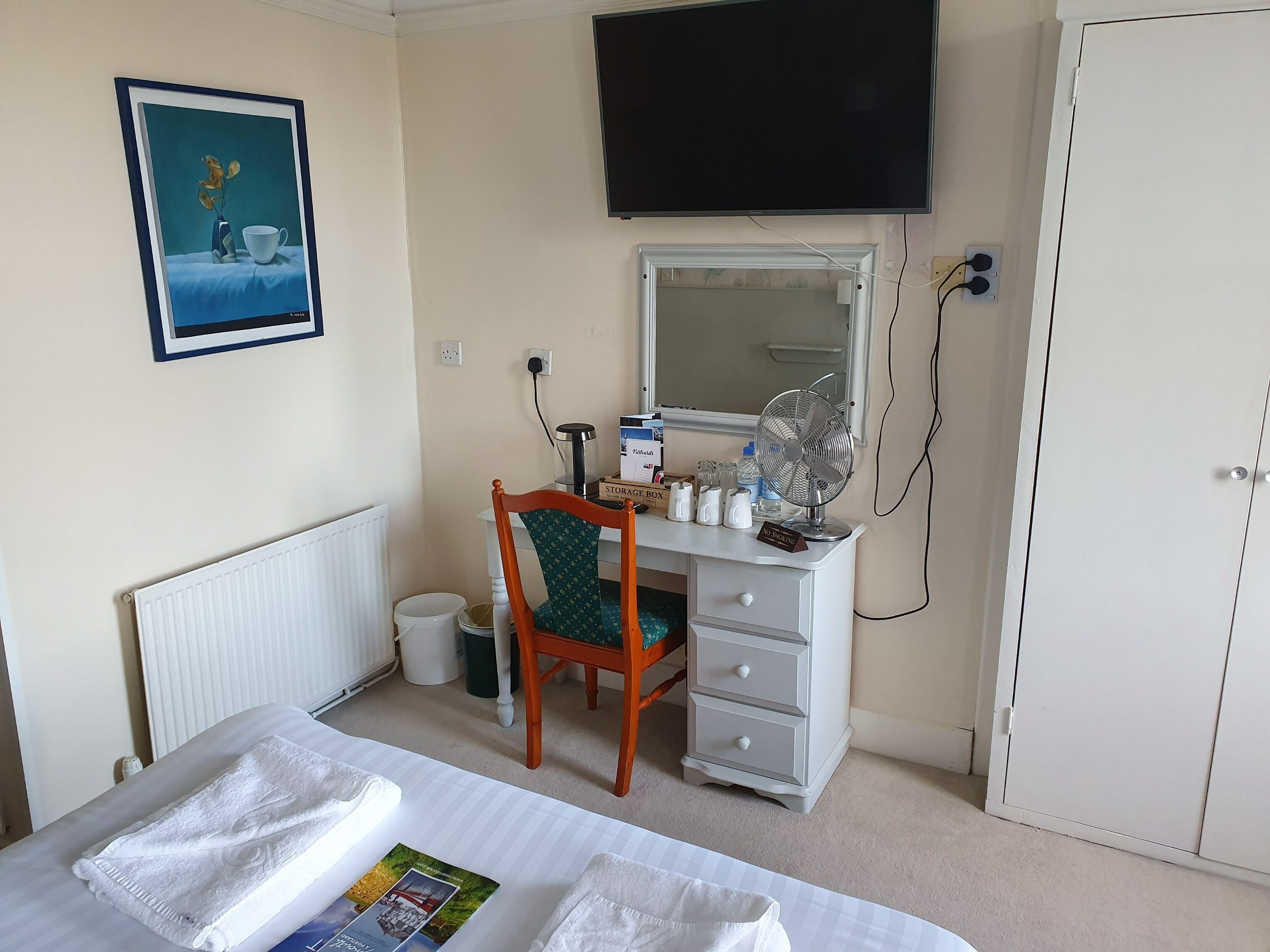 Habitación de hotel con cama, escritorio con silla, televisor, radiador, cubo de basura y armario.