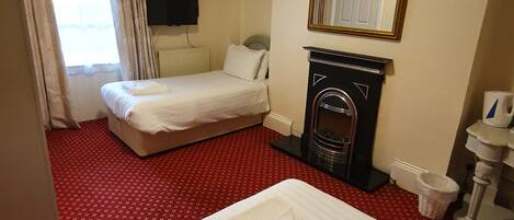 A hotel room with a bed, a small fireplace, a mirror, a radiator, and a window with curtains.