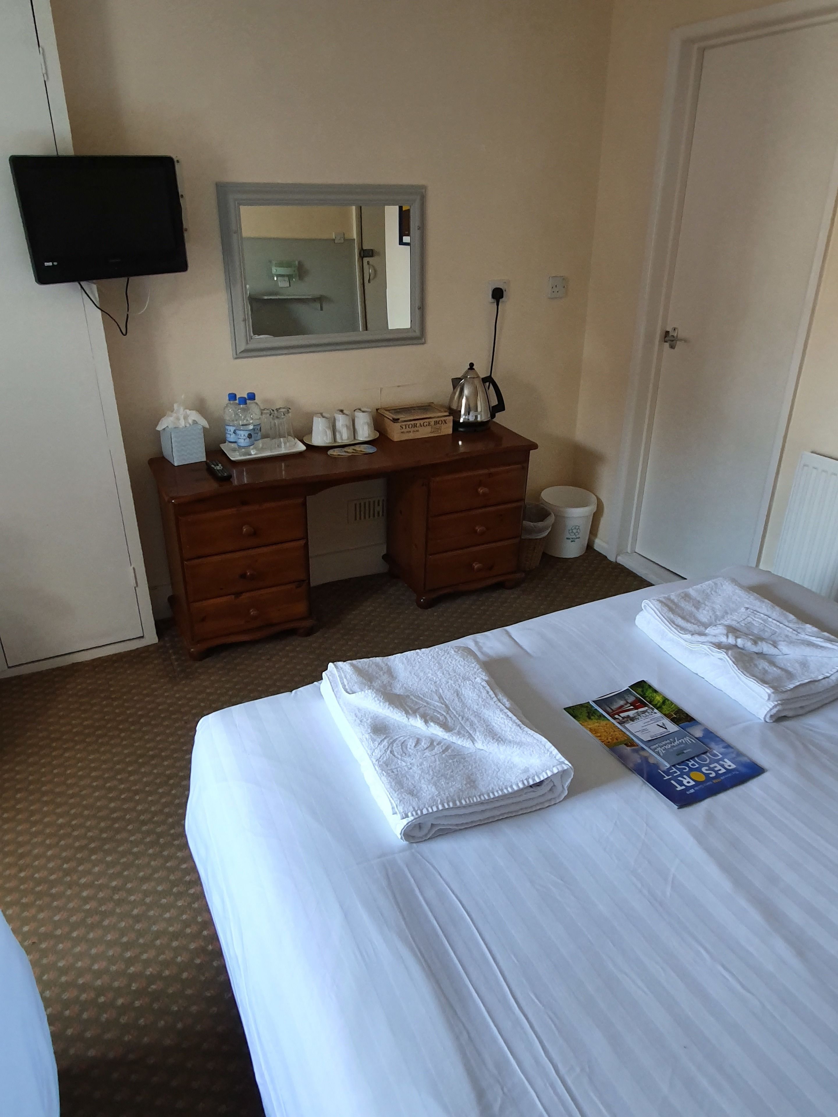 Quarto de hotel com cama, secretária com espelho, caixa de armazenamento e chaleira.