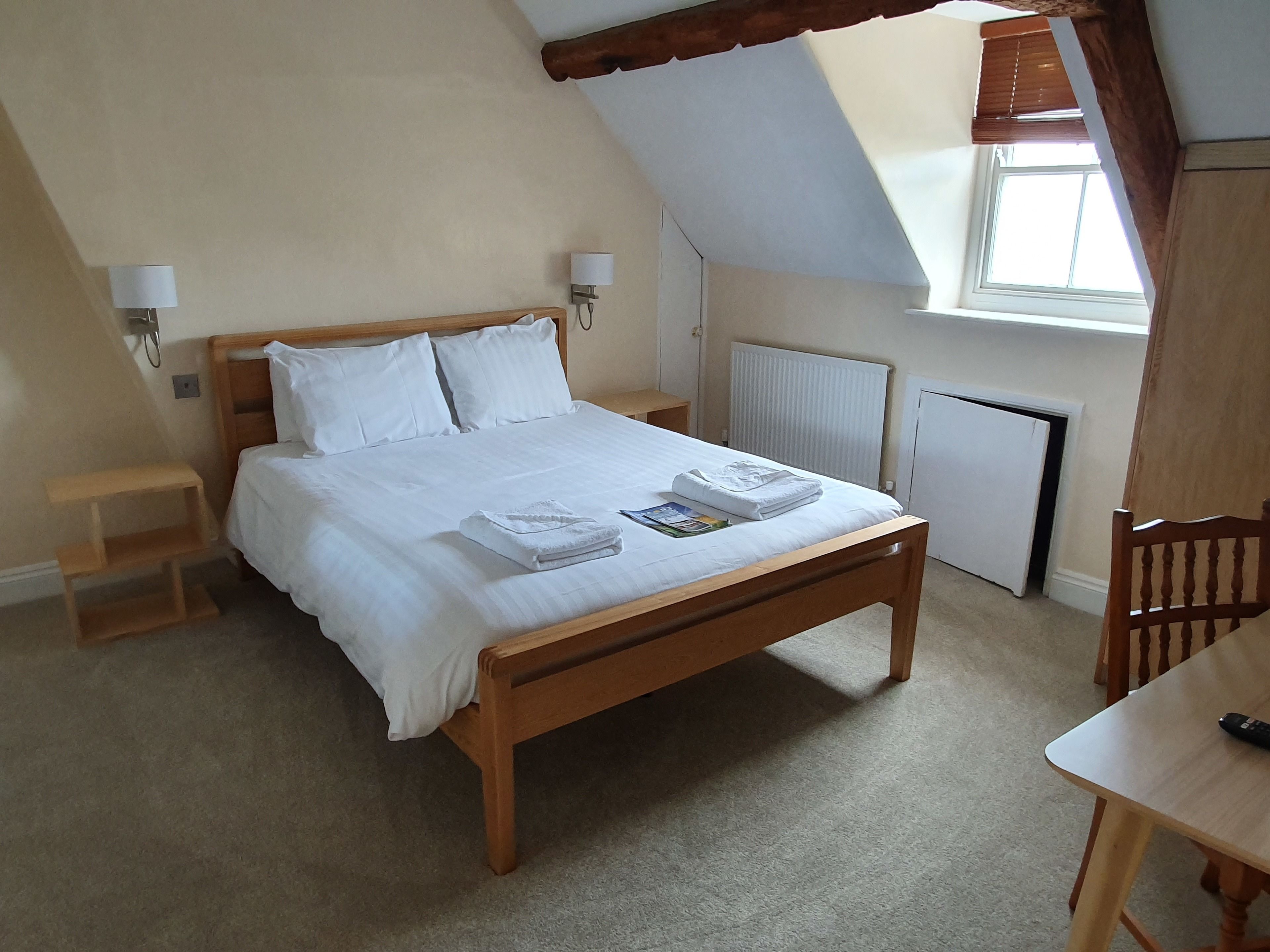 A neatly made bed with white linens and towels, a wooden bedside table, a small wooden stool, and a window with a view of the outside.