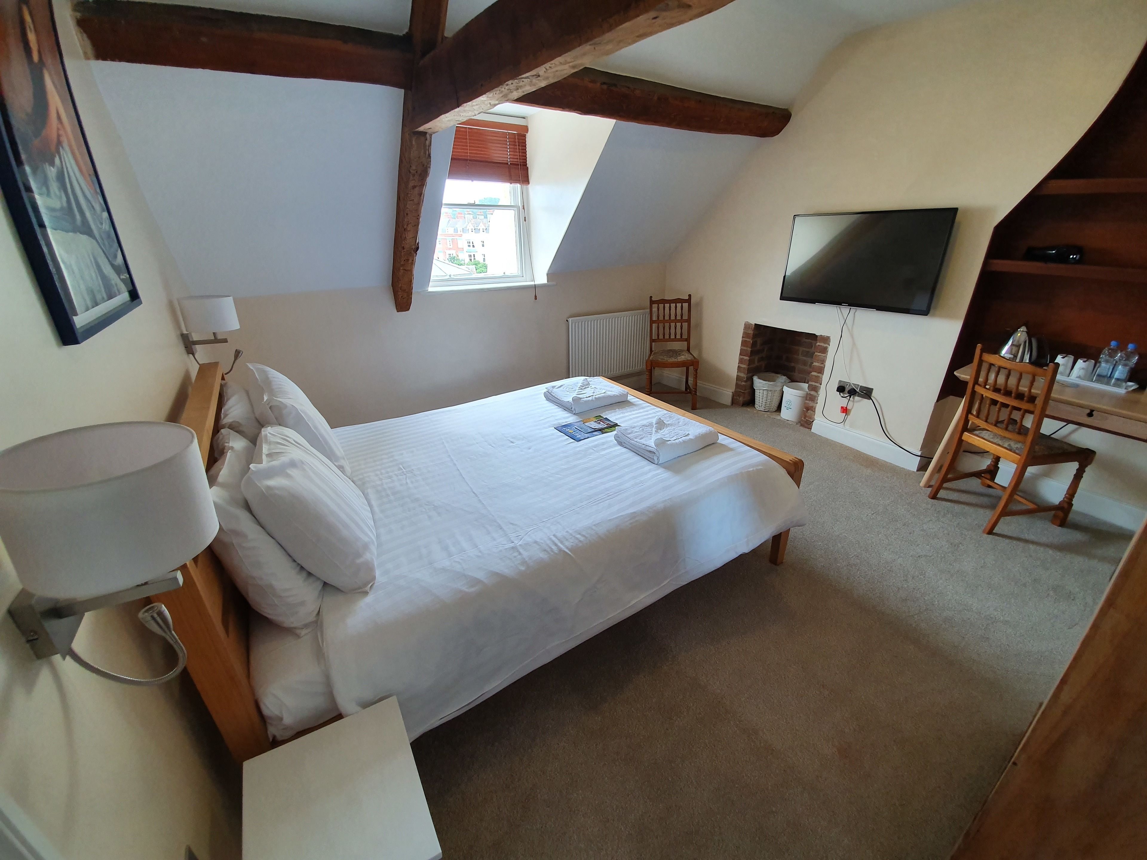 A bedroom with a bed, a desk, a chair, a TV, and a fireplace.