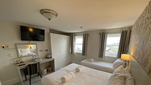 A hotel room with two beds, a desk with a mirror, a television, and a window with curtains. - Alexandra Hotel (Weymouth)