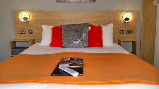 A hotel room with a bed, a painting of a sunset, two bedside lamps, and a remote control on the bed. - The Beaucliffe (Blackpool)