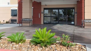 A building entrance with a glass door, two benches, and a small garden area with plants and rocks. - Quality Inn & Suites Kenedy - Karnes City (Kenedy)