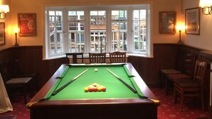 Une table de billard dans une pièce aux poutres apparentes, recouverte d’une moquette rouge, et offrant une vue sur l’extérieur grâce à de grandes fenêtres.