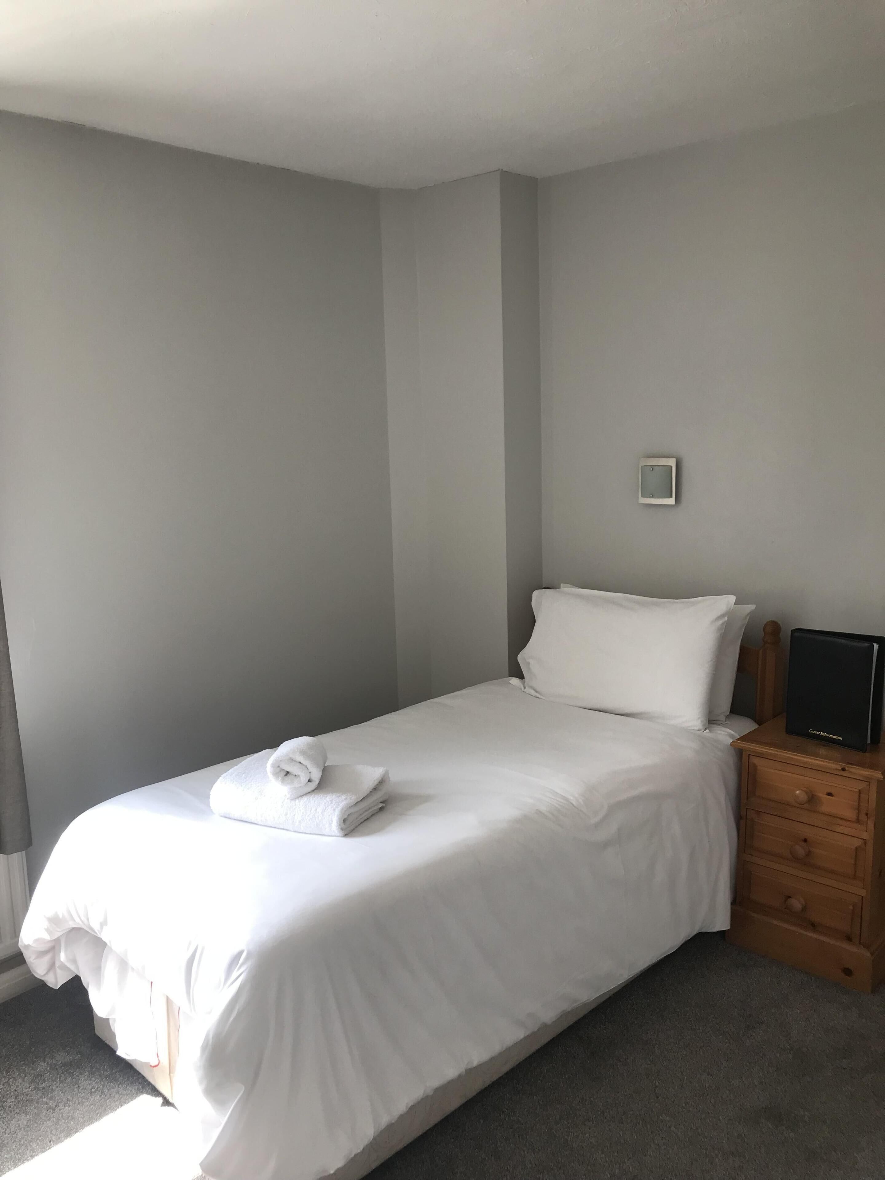 A neatly made bed with a folded towel, a bedside table with a remote control, and a small wall-mounted light fixture.