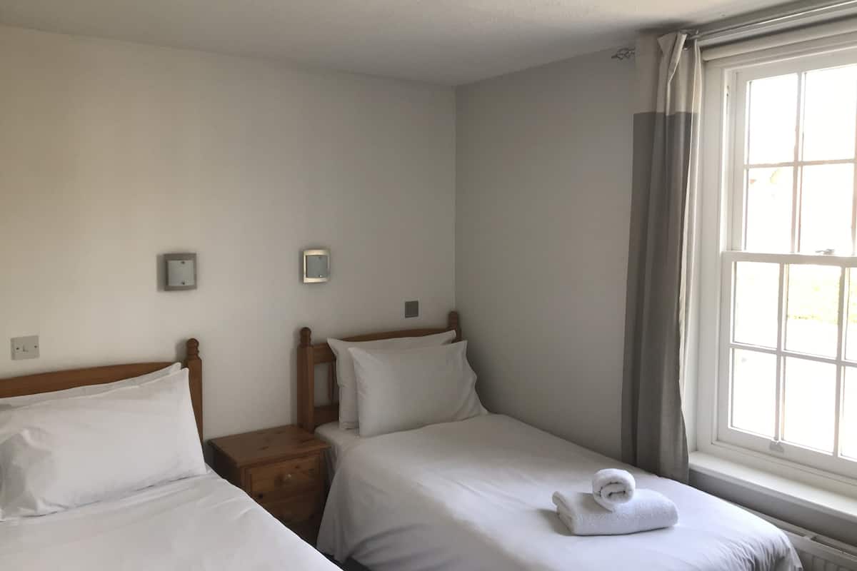 A small, clean room with two single beds, a nightstand, a window with curtains, and a thermostat on the wall.