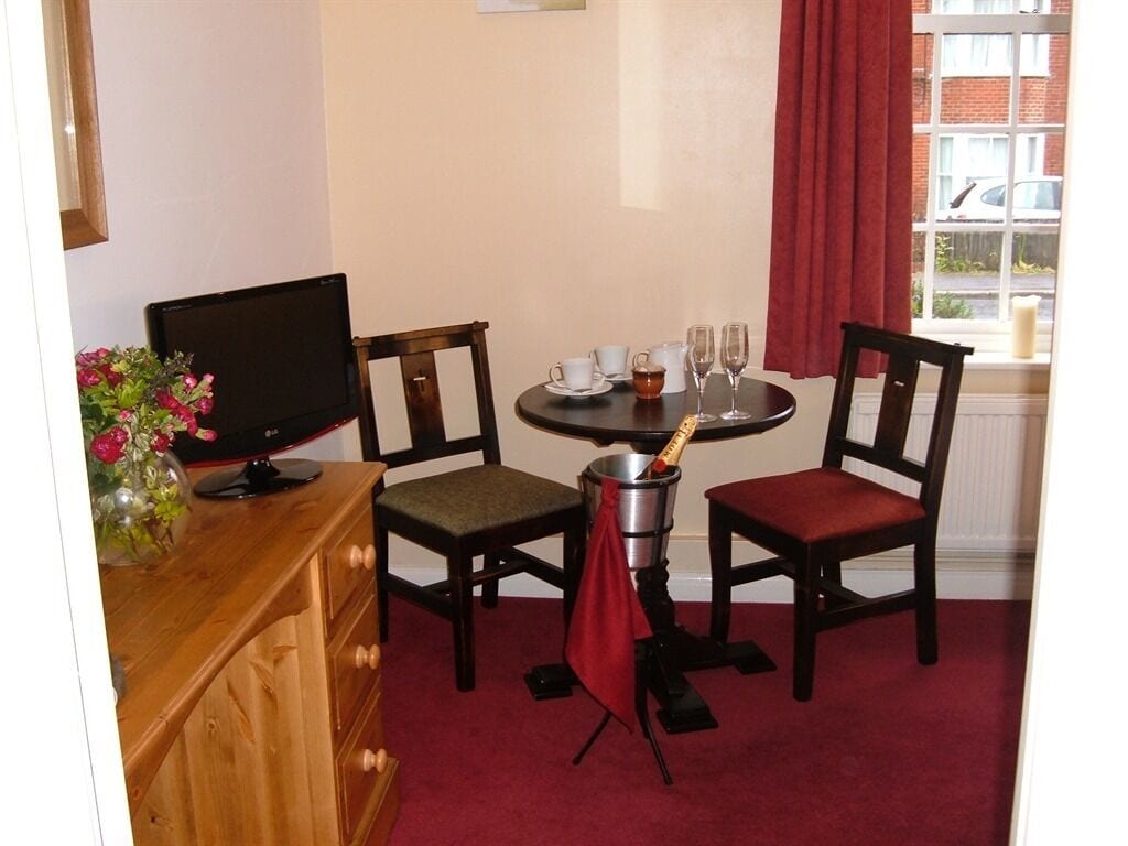 A room with a wooden desk, a television, two chairs, a small table with cups and a bottle, and a window with red curtains.