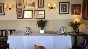 A dining area with a long table set for a meal, framed pictures on the wall, and a small table with a lamp and flowers.