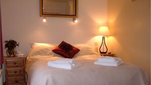 Un lit bien fait, avec du linge de lit blanc et un coussin décoratif rouge, une lampe de chevet et une petite table de nuit en bois dotée de tiroirs.