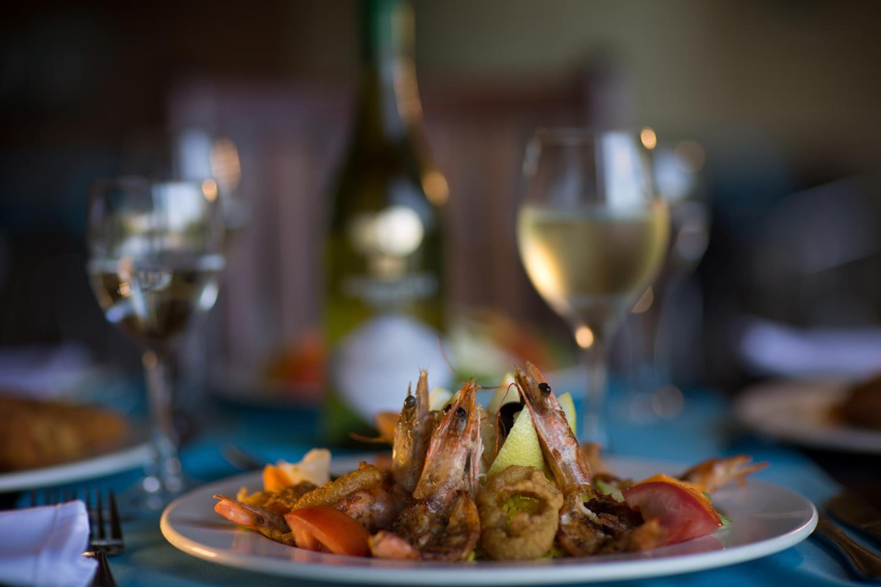Une assiette de fruits de mer, comprenant des crevettes et des noix de Saint-Jacques, servie avec des légumes et accompagnée d’un verre de vin blanc.
