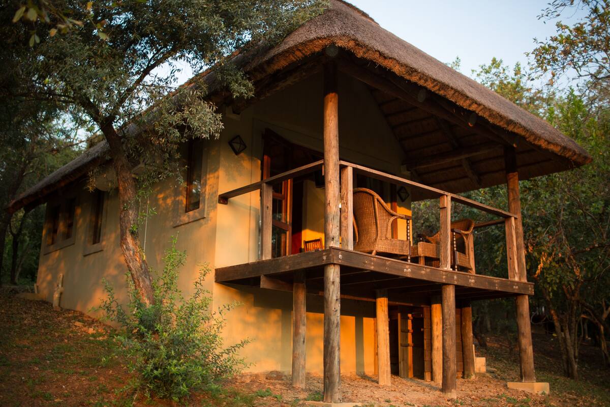 Une structure rustique en bois, dotée d’un toit de chaume, d’un balcon agrémenté de meubles en osier et entourée d’un paysage naturel.