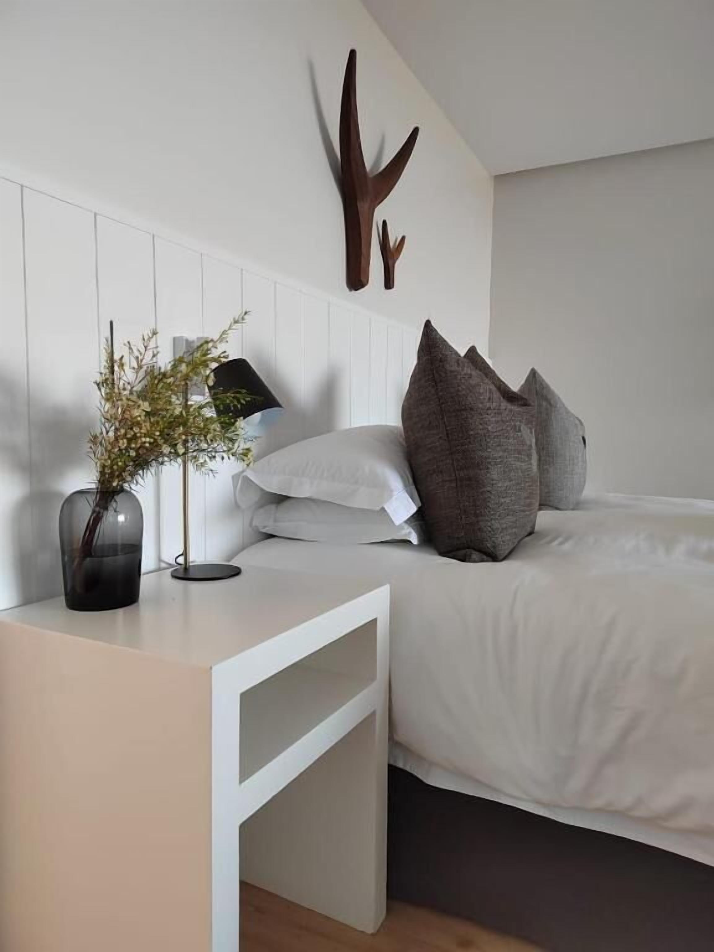 Quarto moderno com cama branca, almofadas cinzas, um vaso com flores secas e um ornamento de chifre de veado fixado na parede.