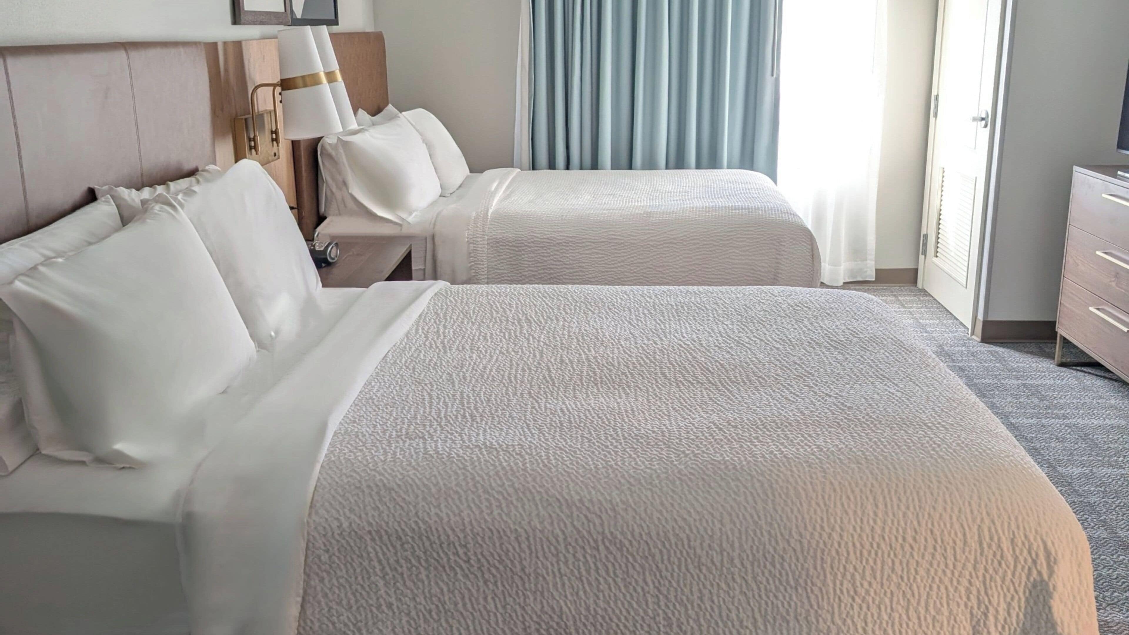 A hotel room with a large bed, two bedside tables, a lamp, and a view of the outside through curtains.