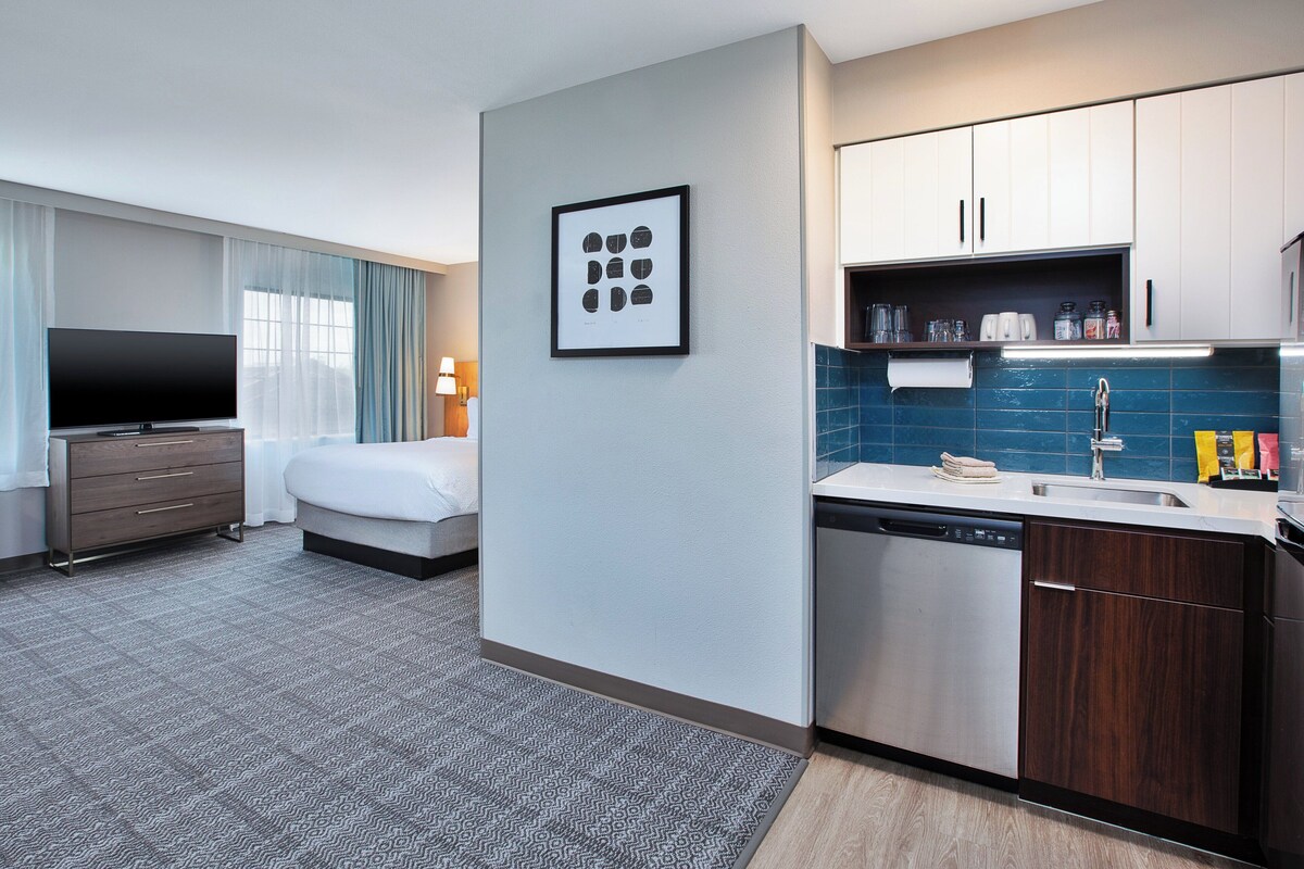 A modern hotel room with a flat-screen TV, a bed, a small kitchenette, and a bathroom.