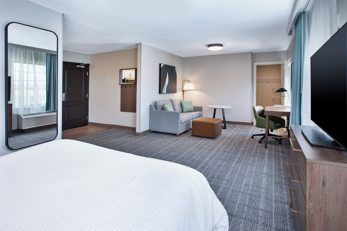 A hotel room with a large bed, a sofa, a desk, a chair, a small table, a mirror, and a television.