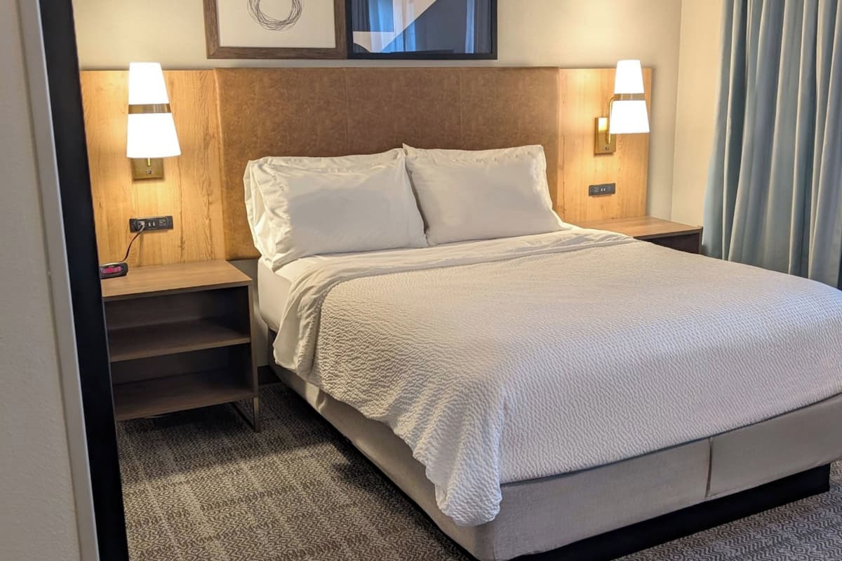 Ein Hotelzimmer mit Bett, zwei Nachttischlampen, einem Nachttisch, einer Steckdose und einem Vorhangfenster.