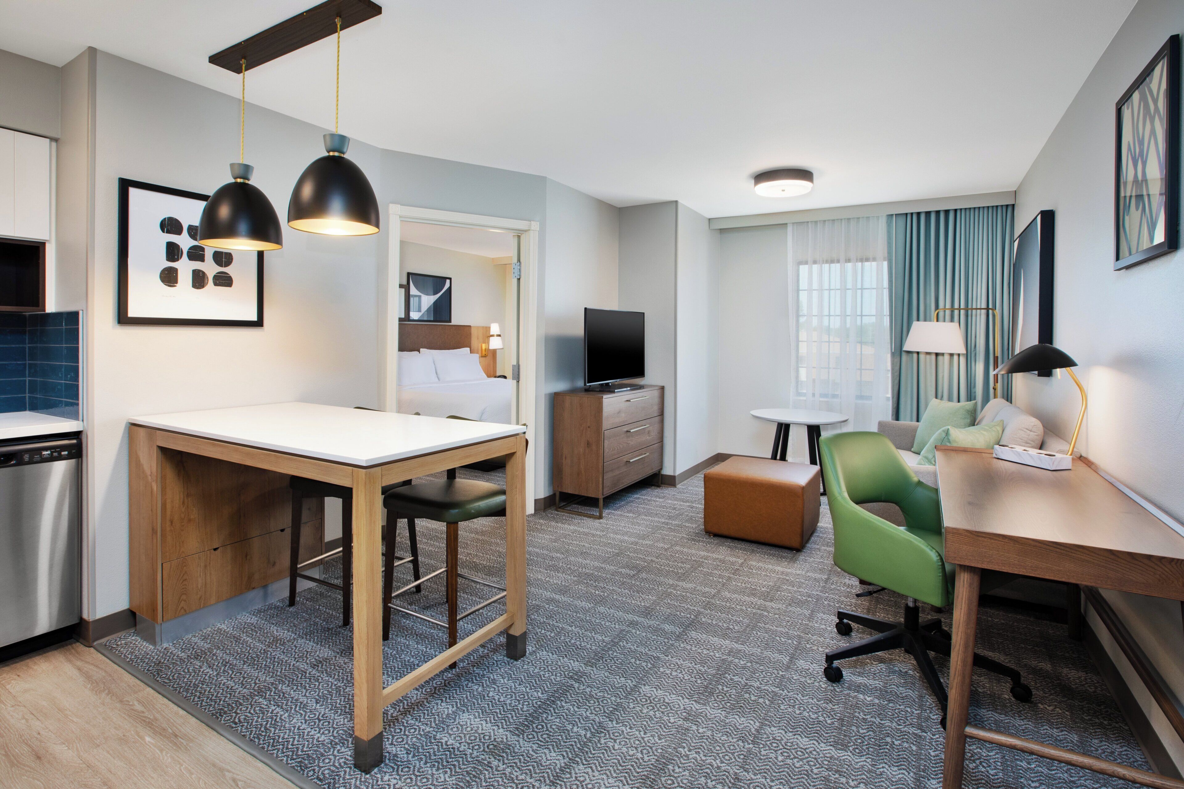 A modern hotel room with a kitchenette, a dining area, a living area with a sofa and armchair, a flat-screen TV, and a large window with curtains.