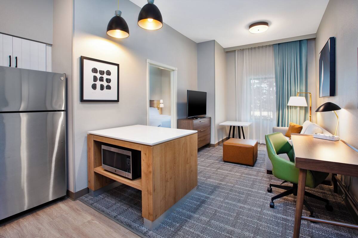 Ein kompaktes Hotelzimmer mit Küchenzeile, einer kleinen Essecke, einer Sitzecke und einem großen Fenster mit Vorhängen.