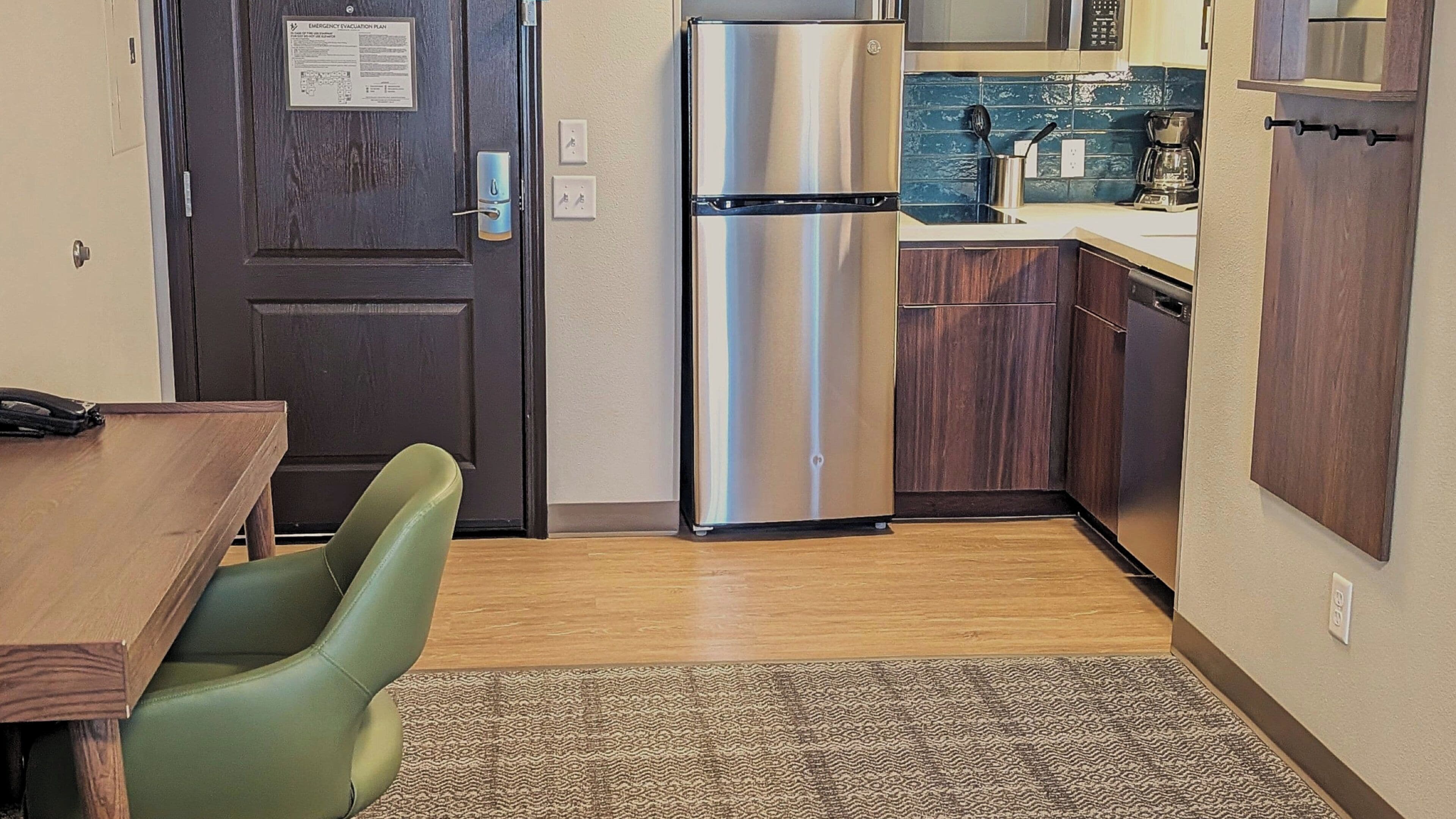 A compact kitchen with a stainless steel refrigerator, wooden cabinets, and a small dining area with a green chair.