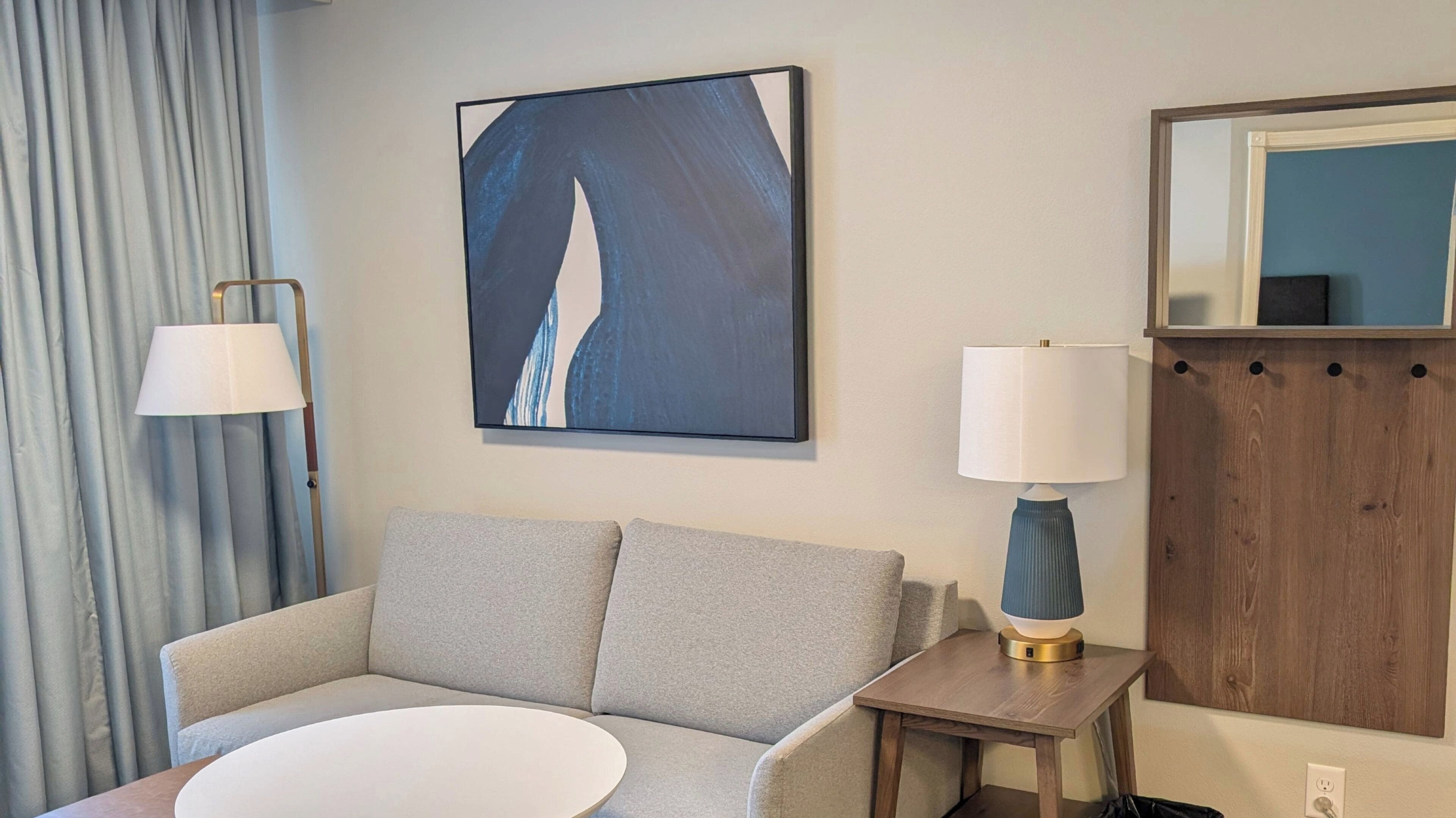 A hotel room with a grey sofa, a round white table, a blue abstract painting, a white lamp, and a wooden cabinet with a mirror.