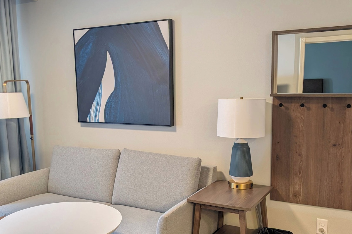 A hotel room with a grey sofa, a round white table, a blue abstract painting, a white lamp, and a wooden cabinet with a mirror.