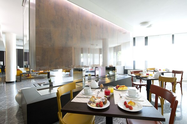 Un espace repas moderne, avec des tables préparées pour le petit-déjeuner, où l’on trouve des assiettes de fruits, des viennoiseries et diverses boissons.
