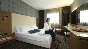 Habitación de hotel con cama, escritorio con una computadora portátil, una silla y una bandeja de desayuno con frutas y pastelería.